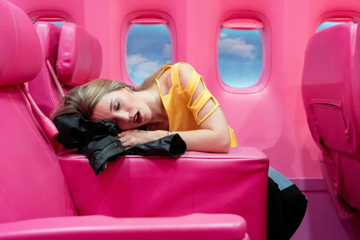 A woman in a yellow top is asleep across bright pink airplane seats in a matching bright pink airplane interior, resting her head on a black jacket.