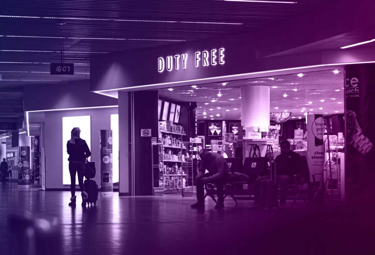 Travellers outside a brightly lit duty free store in an airport terminal, with shelves of products and signage visible inside.