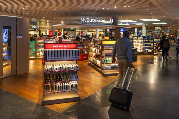 Travellers walk through a brightly lit duty-free store at an airport, passing shelves of discounted perfumes and cosmetics, with prominent sale signage and branded displays.
