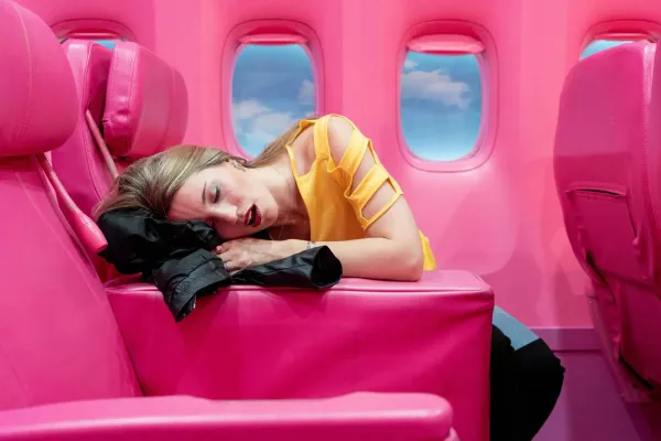 A woman in a yellow top is asleep across bright pink airplane seats in a matching bright pink airplane interior, resting her head on a black jacket.