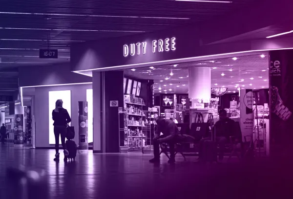 Travellers outside a brightly lit duty free store in an airport terminal, with shelves of products and signage visible inside.
